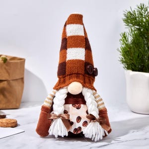 May include: A coffee-themed gnome doll with a brown and white checkered hat, a button detail, and white braided hair. The doll has a beige nose, brown mittens, and a dress with coffee bean print. A decorative item.