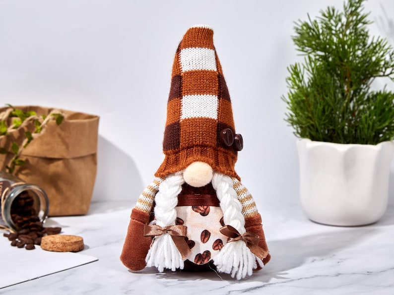 May include: A coffee-themed gnome doll with a brown and white checkered hat, a button detail, and white braided hair. The doll has a beige nose, brown mittens, and a dress with coffee bean print. A decorative item.