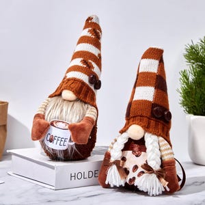 May include: Two coffee-themed gnomes with brown and white striped hats and long white beards. One gnome holds a mug with "Coffee" printed on it. The other gnome has a coffee bean pattern on its body. Decorative items.