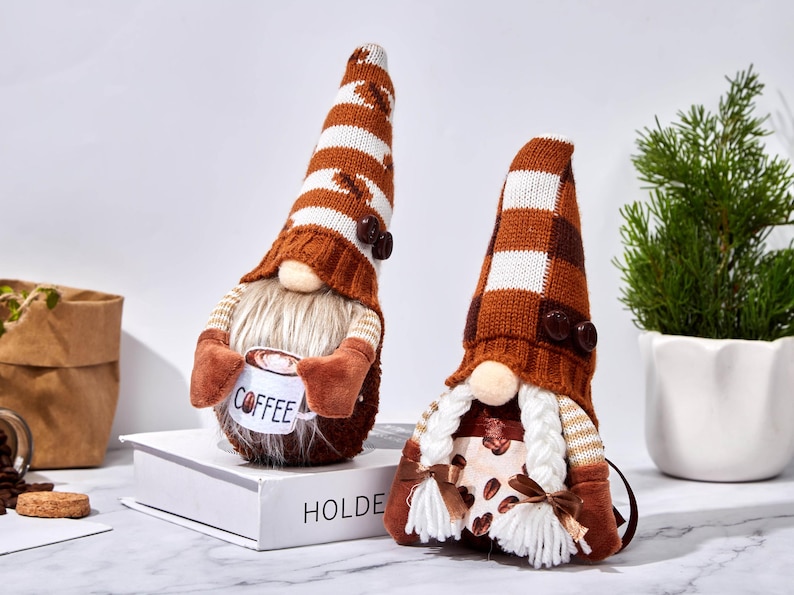 May include: Two coffee-themed gnomes with brown and white striped hats and long white beards. One gnome holds a mug with "Coffee" printed on it. The other gnome has a coffee bean pattern on its body. Decorative items.