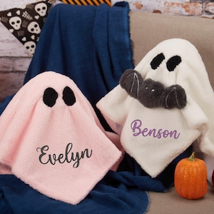 May include: Two plush ghost-shaped blankets, one pink and one white, with black eye cutouts. The pink blanket has the name "Evelyn" embroidered, and the white blanket has "Benson". Halloween-themed decor.