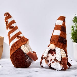 May include: Two decorative gnomes with brown and white knit hats and long white beards. One gnome has a striped hat, the other has a checkered hat. Both gnomes have brown bodies and arms. The gnomes are on a white surface.