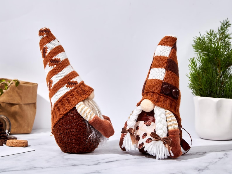 May include: Two decorative gnomes with brown and white knit hats and long white beards. One gnome has a striped hat, the other has a checkered hat. Both gnomes have brown bodies and arms. The gnomes are on a white surface.