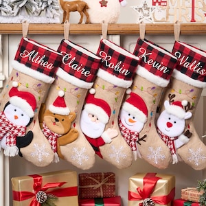May include: Five Christmas stockings with names, featuring festive characters such as a penguin, bear, Santa, snowman, and reindeer. Each stocking has a red and black plaid cuff and is decorated with snowflakes and Christmas lights.