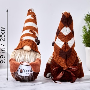 May include: Two decorative gnomes, each approximately 9.9 inches (25 cm) tall. One gnome holds a coffee cup. They have brown and white striped hats, beige beards, and brown mittens. The gnomes are a charming addition to home decor.