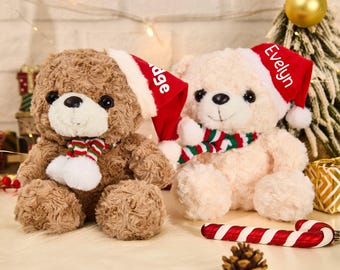 May include: Two plush teddy bears, one brown and one cream-colored, are dressed for the holidays. Each bear wears a red Santa hat with a name on it and a striped scarf. A candy cane and pinecone are in the foreground.