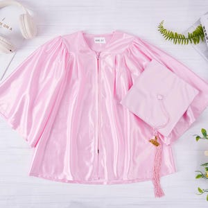 May include: Pink graduation gown and cap set. The gown has a zipper closure and the cap has a tassel.