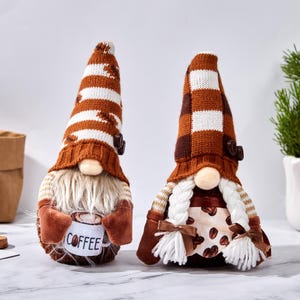 May include: Two coffee-themed gnomes with long, striped hats in brown and white. One gnome holds a mug with "COFFEE" printed on it. The other gnome has braided hair and a dress with coffee bean details. They are decorative items.