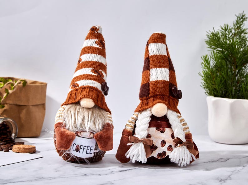May include: Two coffee-themed gnomes with long, striped hats in brown and white. One gnome holds a mug with "COFFEE" printed on it. The other gnome has braided hair and a dress with coffee bean details. They are decorative items.