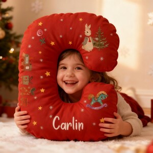 Op de afbeelding: Een rood kerstkussen in de vorm van de letter "C", versierd met geborduurde sterren en feestelijke motieven. Het kussen heeft een konijn en een schommelpaard ontwerp, met de naam "Carlin" erop.