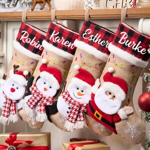 May include: Four Christmas stockings, each personalised with a name: Robin, Karen, Esther, and Burke. They have a burlap design with red and black plaid cuffs. Festive characters decorate the stockings: a reindeer, penguin, snowman, and Santa Claus.