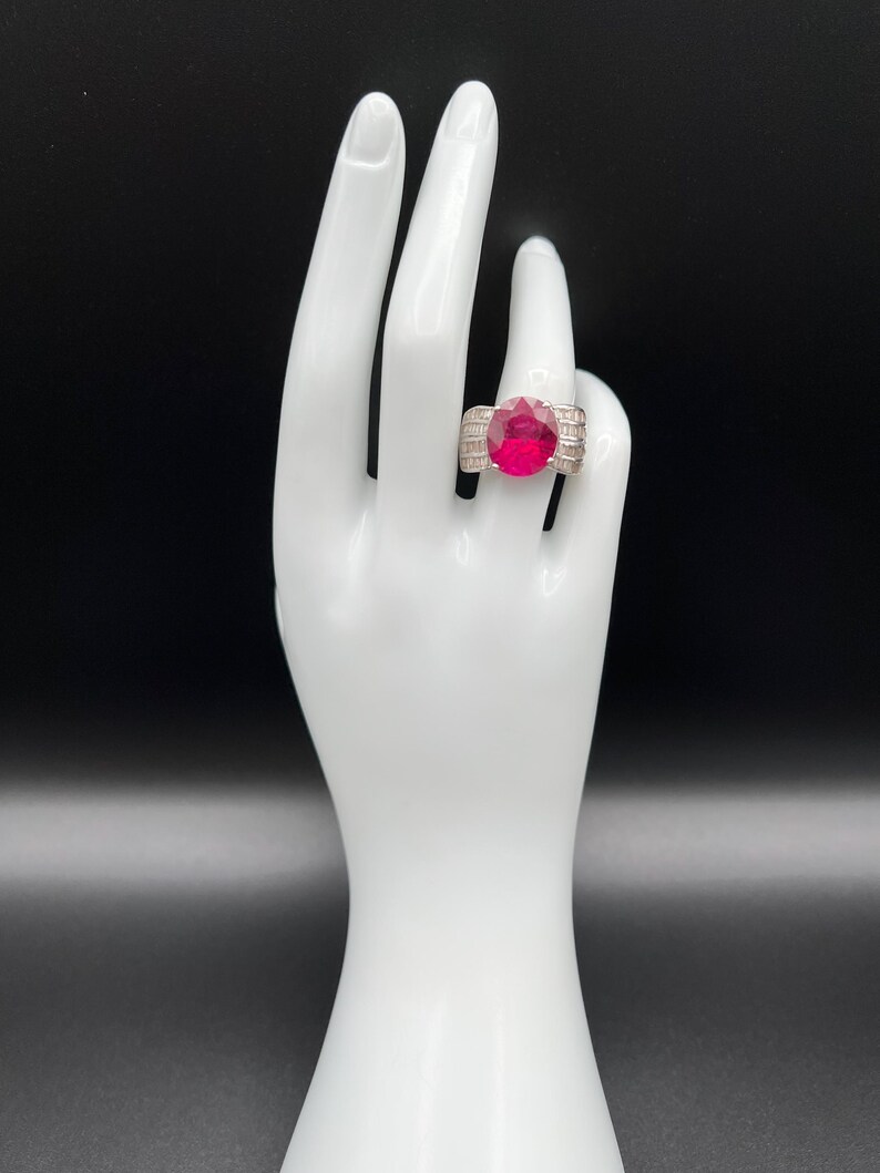 May include: A silver-toned ring featuring a large, oval-cut vibrant red center stone. The band is accented with multiple rows of small, clear baguette-cut stones arranged horizontally along the sides, creating a wide, structured appearance.