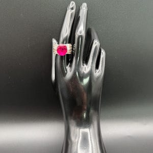 May include: A silver-toned ring featuring a large, oval-cut vibrant pink center stone. The band is wide and constructed with multiple horizontal metallic ridges that extend from the central setting, creating a structured and polished appearance.