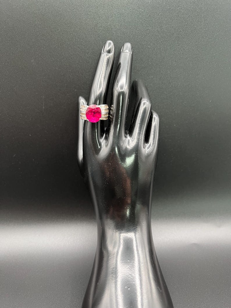 May include: A silver-toned ring featuring a large, oval-cut vibrant pink center stone. The band is wide and constructed with multiple horizontal metallic ridges that extend from the central setting, creating a structured and polished appearance.