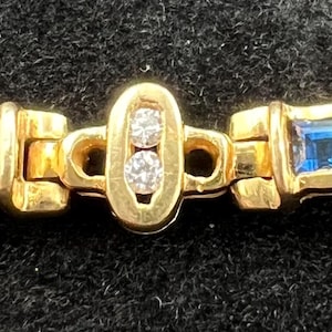 May include: A gold bracelet featuring two rectangular segments inlaid with square-cut blue stones. The segments are connected by a central oval link containing two small, round, clear stones set vertically within the gold frame.