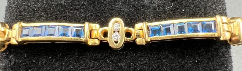 May include: A gold bracelet featuring two rectangular segments inlaid with square-cut blue stones. The segments are connected by a central oval link containing two small, round, clear stones set vertically within the gold frame.