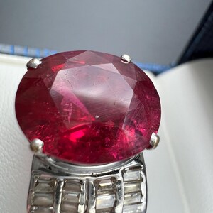 May include: A ring featuring a large, oval-cut, deep red gemstone set in a silver-toned metal band. The band is decorated with multiple small, rectangular, clear stones arranged in rows along the sides, secured by four prominent metal prongs.