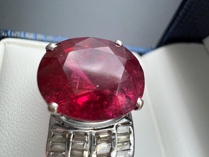 May include: A ring featuring a large, oval-cut, deep red gemstone set in a silver-toned metal band. The band is decorated with multiple small, rectangular, clear stones arranged in rows along the sides, secured by four prominent metal prongs.