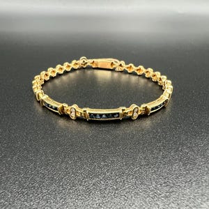 May include: A gold-toned link bracelet featuring a series of small, dark blue rectangular stones set in a channel style. The bracelet includes decorative metal accents between the stone segments and a secure clasp mechanism at the top center.
