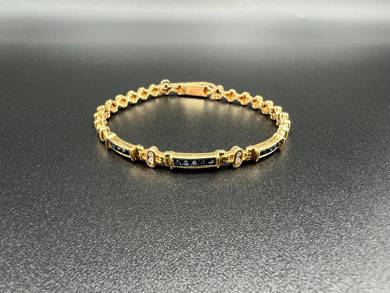 May include: A gold-toned link bracelet featuring a series of small, dark blue rectangular stones set in a channel style. The bracelet includes decorative metal accents between the stone segments and a secure clasp mechanism at the top center.
