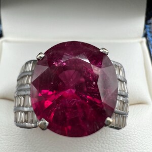 May include: A ring featuring a large, oval-cut, deep magenta gemstone set in a silver-toned metal band. The band is accented with rows of small, clear, baguette-cut stones on both sides, secured by four metal prongs holding the central stone.