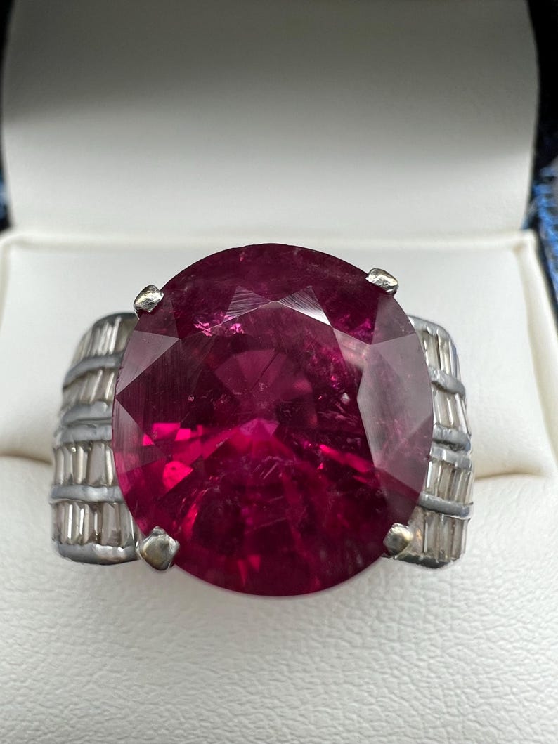 May include: A ring featuring a large, oval-cut, deep magenta gemstone set in a silver-toned metal band. The band is accented with rows of small, clear, baguette-cut stones on both sides, secured by four metal prongs holding the central stone.