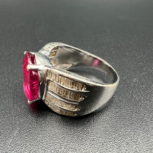 May include: A silver-toned ring featuring a large, rectangular, vibrant pink center stone. The band is wide and decorated with three rows of small, clear, rectangular baguette-cut stones set along the sides, creating a faceted, reflective appearance.