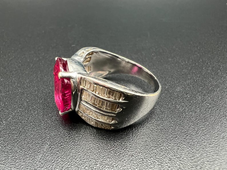 May include: A silver-toned ring featuring a large, rectangular, vibrant pink center stone. The band is wide and decorated with three rows of small, clear, rectangular baguette-cut stones set along the sides, creating a faceted, reflective appearance.