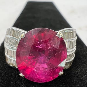 May include: A silver ring featuring a large, round, faceted deep pink gemstone as the centerpiece. The band is wide and decorated with multiple rows of small, clear, rectangular baguette-cut stones set horizontally along the sides of the ring.
