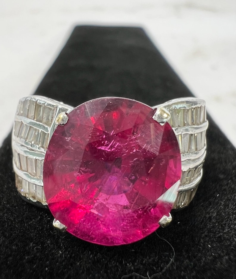 May include: A silver ring featuring a large, round, faceted deep pink gemstone as the centerpiece. The band is wide and decorated with multiple rows of small, clear, rectangular baguette-cut stones set horizontally along the sides of the ring.