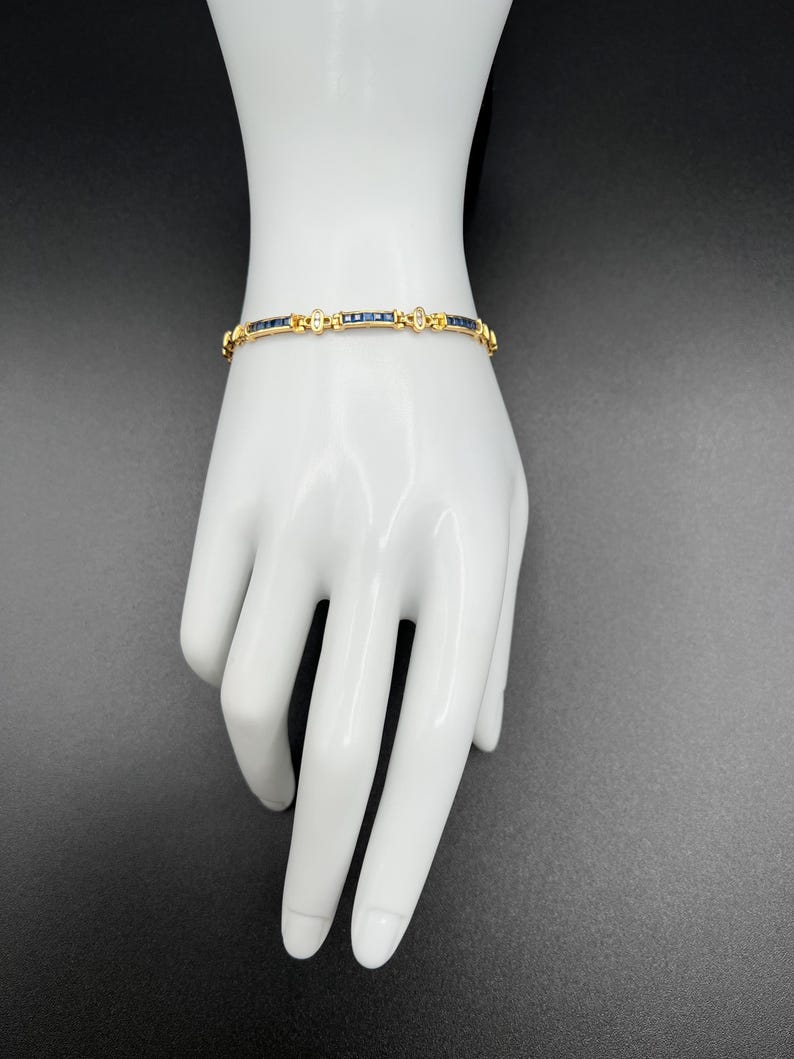 May include: A gold bracelet featuring a series of square-cut dark blue sapphires set in a channel style, interspersed with small, clear, round-cut accent stones. The metal links are polished gold, creating a continuous, flexible band design.