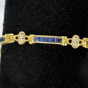 May include: A gold-toned link bracelet featuring alternating segments of channel-set square blue stones and oval-shaped links containing two small round clear stones. The design consists of repeating rectangular sections connected by hinged metal joints.