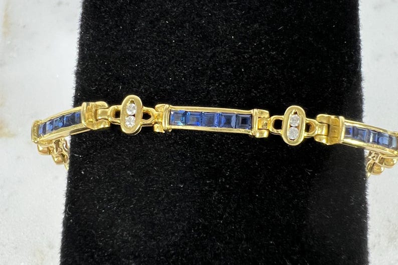 May include: A gold-toned link bracelet featuring alternating segments of channel-set square blue stones and oval-shaped links containing two small round clear stones. The design consists of repeating rectangular sections connected by hinged metal joints.
