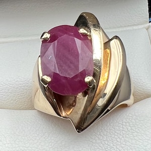 May include: A gold ring featuring a large, oval-cut, deep pinkish-red gemstone held in place by four prongs. The band is constructed from polished yellow gold with a wide, sculptural, and layered design that wraps around the central stone.