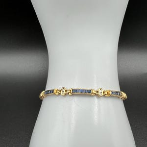 May include: A gold-toned link bracelet featuring a series of rectangular dark blue sapphire-colored stones set in a channel, alternating with small clear stones and gold metal spacers that connect each segment in a continuous chain design.