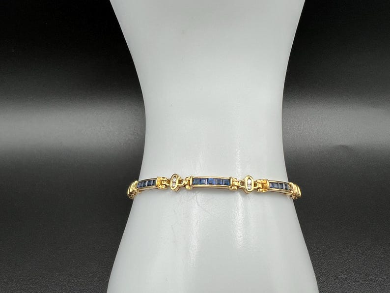 May include: A gold-toned link bracelet featuring a series of rectangular dark blue sapphire-colored stones set in a channel, alternating with small clear stones and gold metal spacers that connect each segment in a continuous chain design.