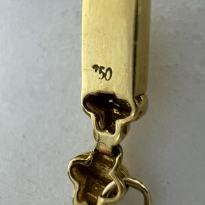 May include: A gold jewelry clasp featuring a rectangular end piece stamped with the number 750. The clasp mechanism includes a hinged component with a decorative, clover-like shape and a small circular jump ring attached to the bottom section.