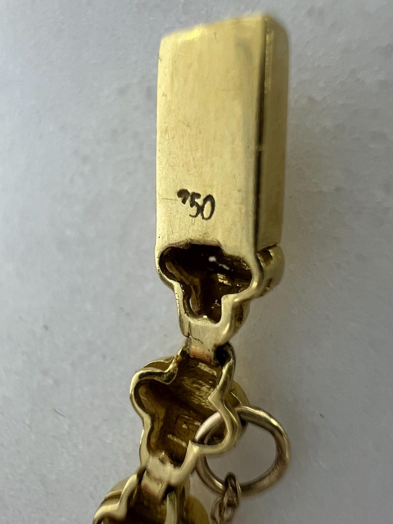 May include: A gold jewelry clasp featuring a rectangular end piece stamped with the number 750. The clasp mechanism includes a hinged component with a decorative, clover-like shape and a small circular jump ring attached to the bottom section.