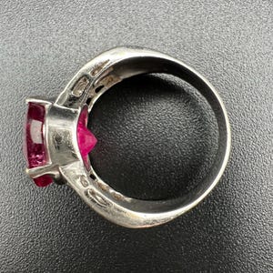May include: A silver-toned ring featuring a prominent, faceted, deep pink gemstone set in a prong setting. The metal band has a smooth, polished finish and a wide profile, showcasing a clear view of the stone's pointed pavilion and the setting.