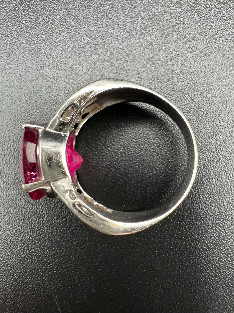 May include: A silver-toned ring featuring a prominent, faceted, deep pink gemstone set in a prong setting. The metal band has a smooth, polished finish and a wide profile, showcasing a clear view of the stone's pointed pavilion and the setting.