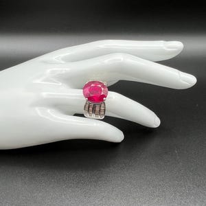 May include: A silver ring featuring a large, round, faceted deep pink gemstone as the centerpiece. The band is wide and constructed from multiple vertical rows of small, clear, rectangular baguette-cut stones set in a polished silver metal frame.