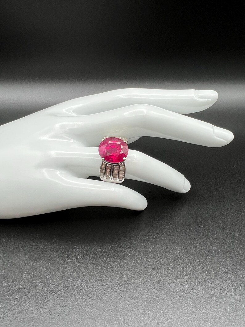 May include: A silver ring featuring a large, round, faceted deep pink gemstone as the centerpiece. The band is wide and constructed from multiple vertical rows of small, clear, rectangular baguette-cut stones set in a polished silver metal frame.