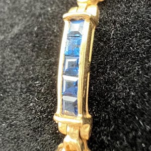 May include: A gold bracelet featuring a series of square-cut blue gemstones set in a channel. The metal links are polished and curved, alternating between sections of blue stones and solid gold segments, creating a continuous, articulated design.