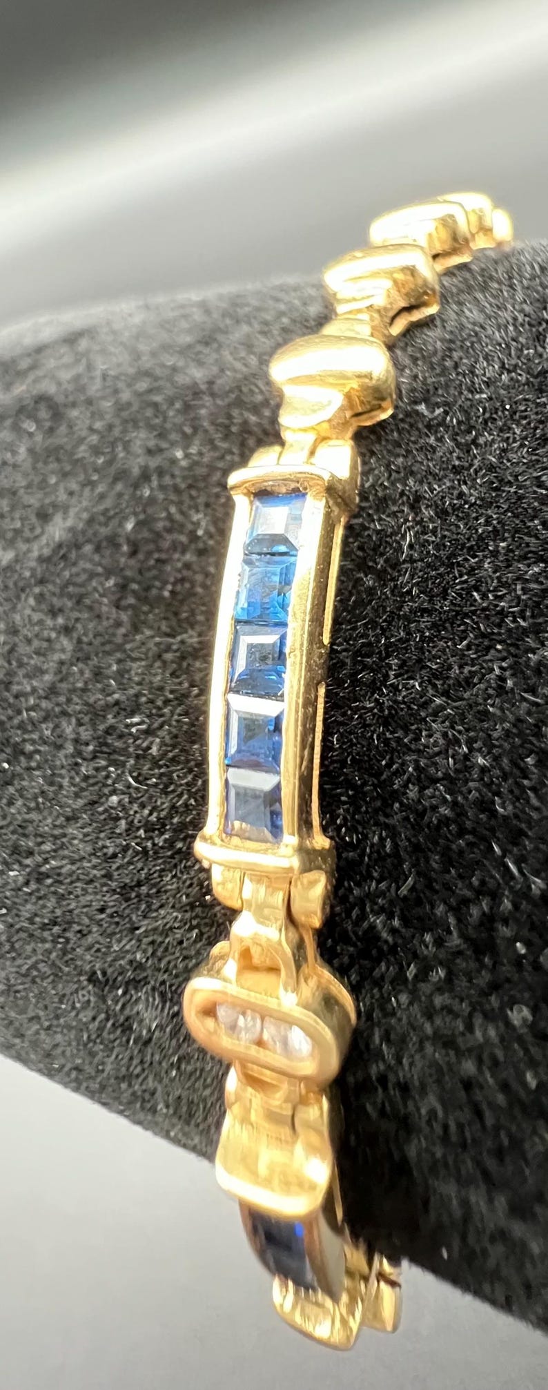 May include: A gold bracelet featuring a series of square-cut blue gemstones set in a channel. The metal links are polished and curved, alternating between sections of blue stones and solid gold segments, creating a continuous, articulated design.