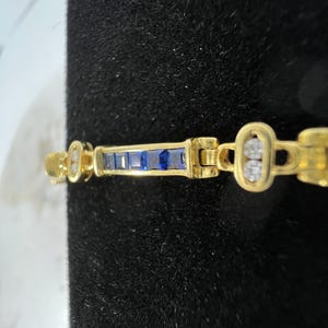 May include: A gold bracelet featuring a central bar set with five rectangular blue sapphires, flanked by oval links each containing two round white diamonds. The metal has a polished finish, and the design consists of alternating sapphire and diamond sections.