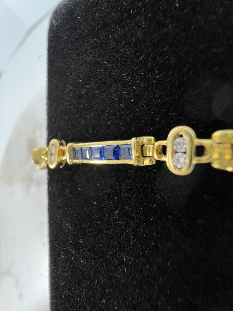 May include: A gold bracelet featuring a central bar set with five rectangular blue sapphires, flanked by oval links each containing two round white diamonds. The metal has a polished finish, and the design consists of alternating sapphire and diamond sections.