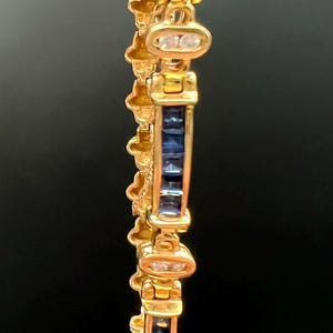May include: A gold bracelet featuring a repeating pattern of rectangular channel-set blue sapphires alternating with oval links containing two small clear stones. The metal has a polished finish and the design includes decorative side elements.