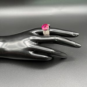 May include: A silver ring featuring a large, round, vibrant pink gemstone set in the center. The band is wide and encrusted with multiple rows of small, clear, rectangular-cut stones that extend along the sides of the metal setting.