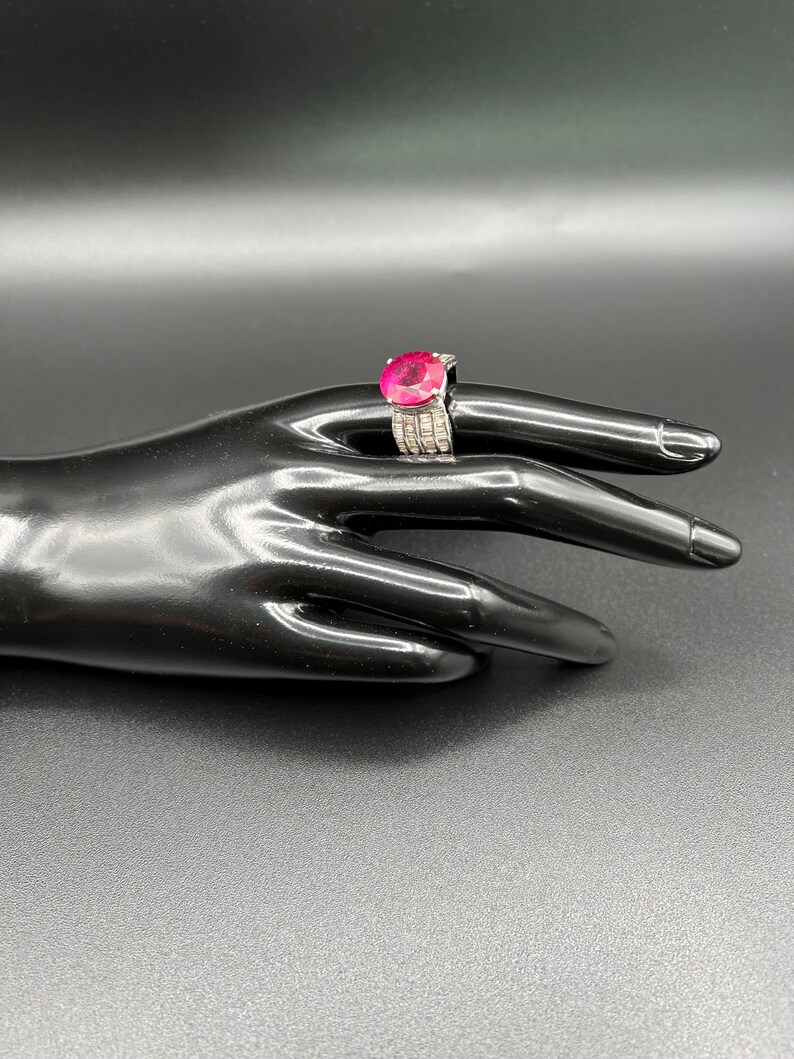 May include: A silver ring featuring a large, round, vibrant pink gemstone set in the center. The band is wide and encrusted with multiple rows of small, clear, rectangular-cut stones that extend along the sides of the metal setting.