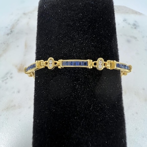 May include: A gold link bracelet featuring a series of square-cut blue gemstones set in a channel style, interspersed with small oval-shaped gold links that each contain two tiny round clear stones arranged vertically along the center.